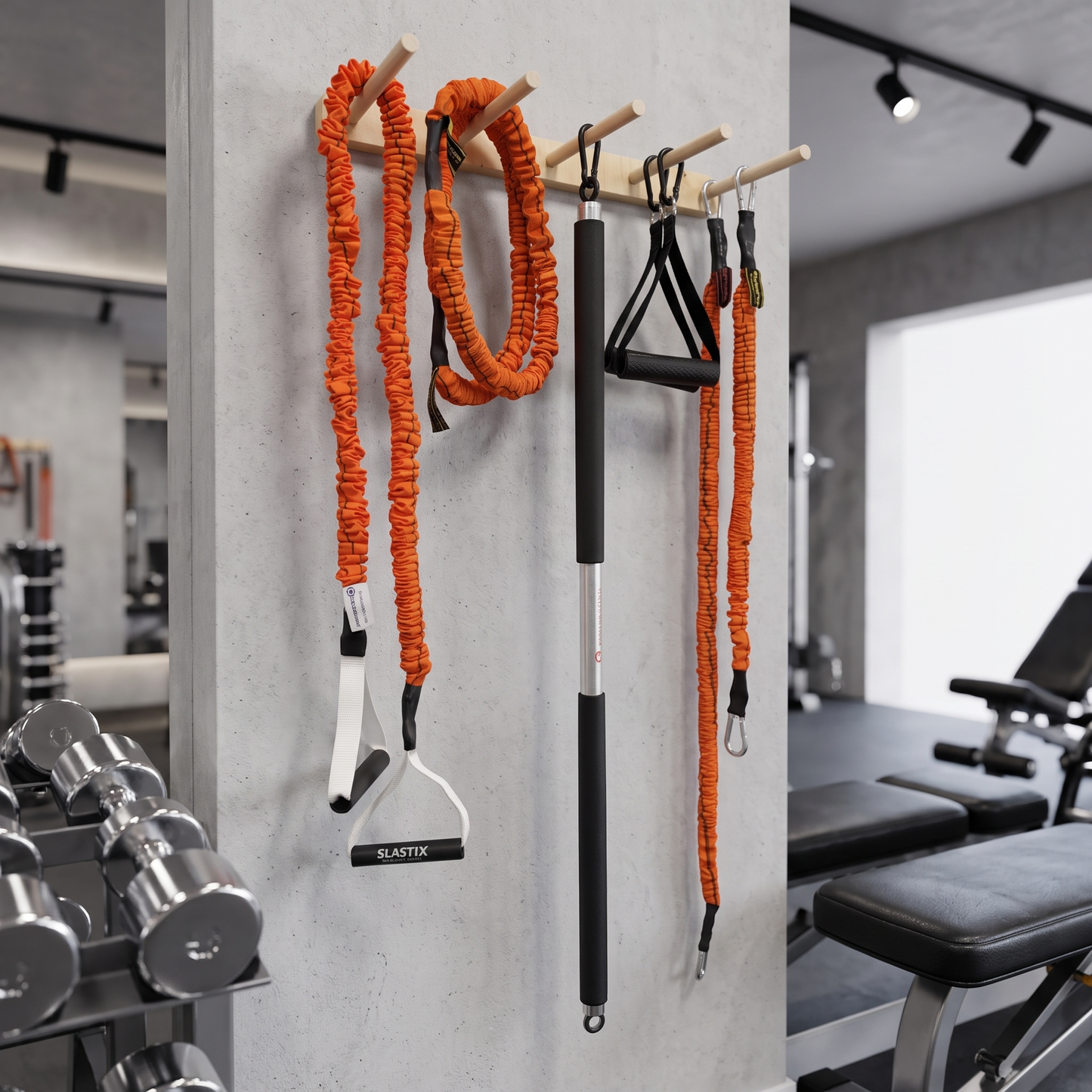 Orange and black resistance bands and a pull-up bar set hanging on a wall rack in a gym, with dumbbells and exercise equipment visible in the background.