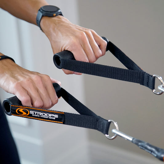 Close-up of a person's hands using a black Stroops dual-pull rope exercise machine, showing the textured handles and metal D-rings against a neutral indoor background.