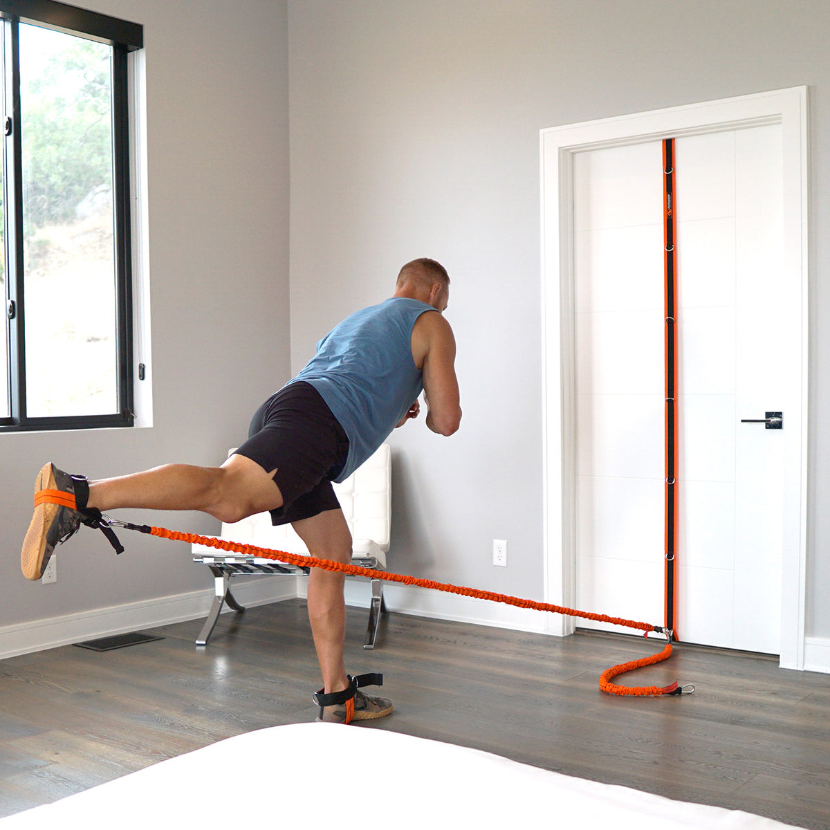A man in a blue tank top and black shorts exercises with an orange resistance band attached to a white door in a modern room with gray walls and wood floors.