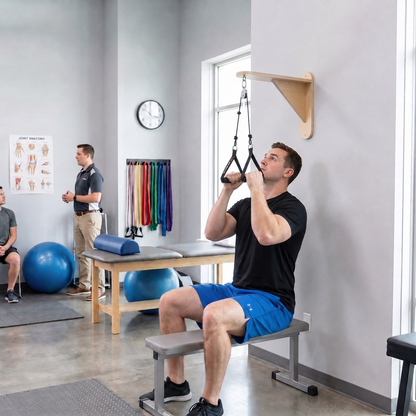 A man in a black t-shirt and blue shorts sits on a bench in a fitness studio, pulling a wall-mounted hanging trapeze bar. Another man stands in the background near exercise equipment.