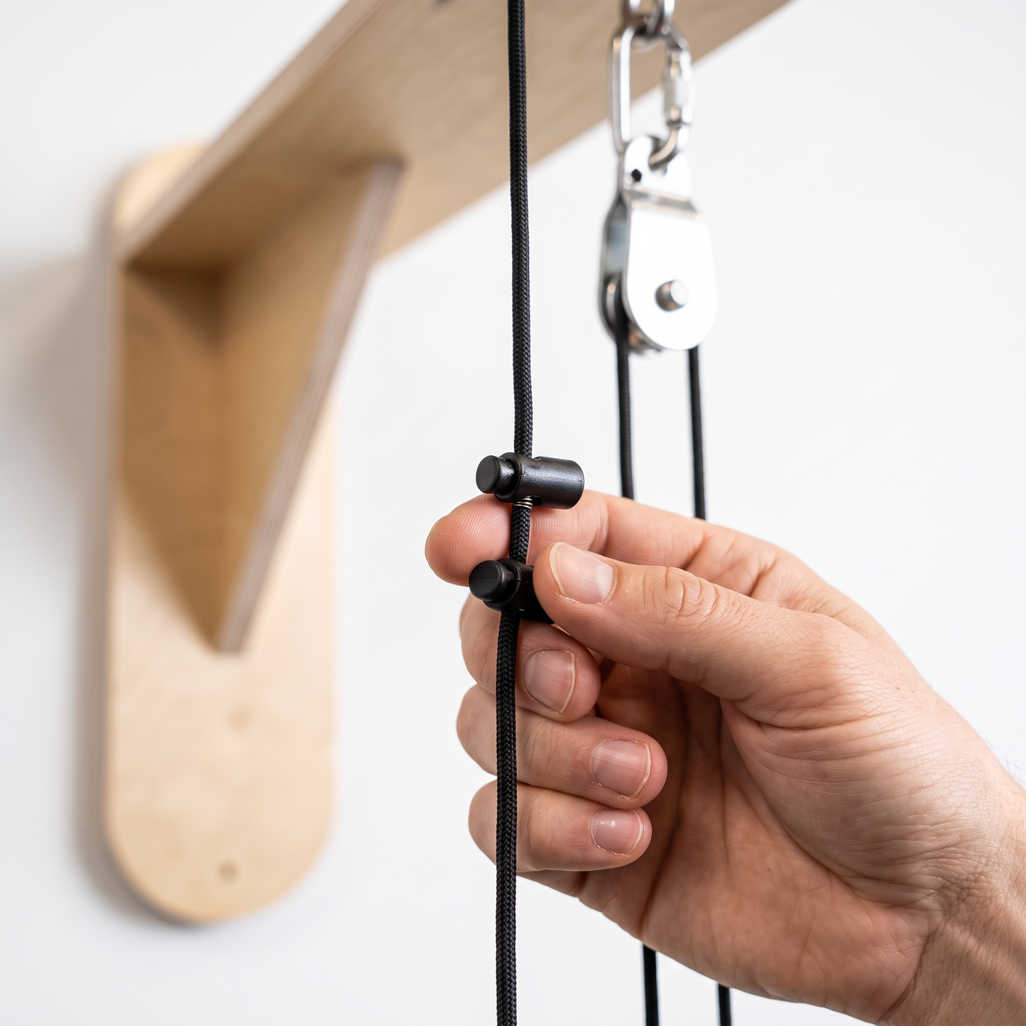 A close-up of a hand adjusting a black cord with a black plastic slider, attached to a pulley system on a wooden wall mount.