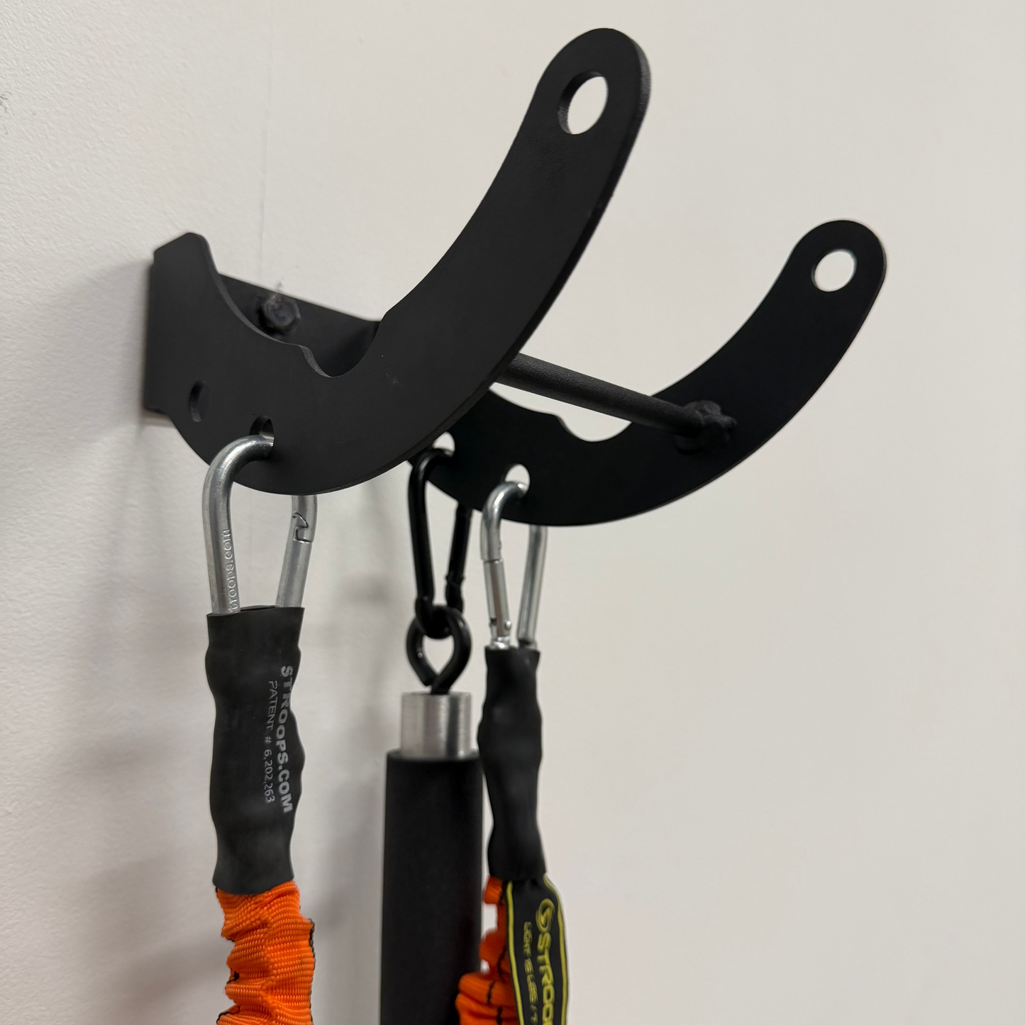 A black metal wall-mounted dual hook rack holding two resistance bands with orange and black handles against a plain off-white wall.
