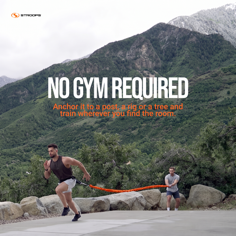 Outdoor promotional scene with two people using an orange battle rope exercise band on a paved path, with large text reading 'NO GYM REQUIRED' and smaller orange copy below. The background shows a mountain landscape.