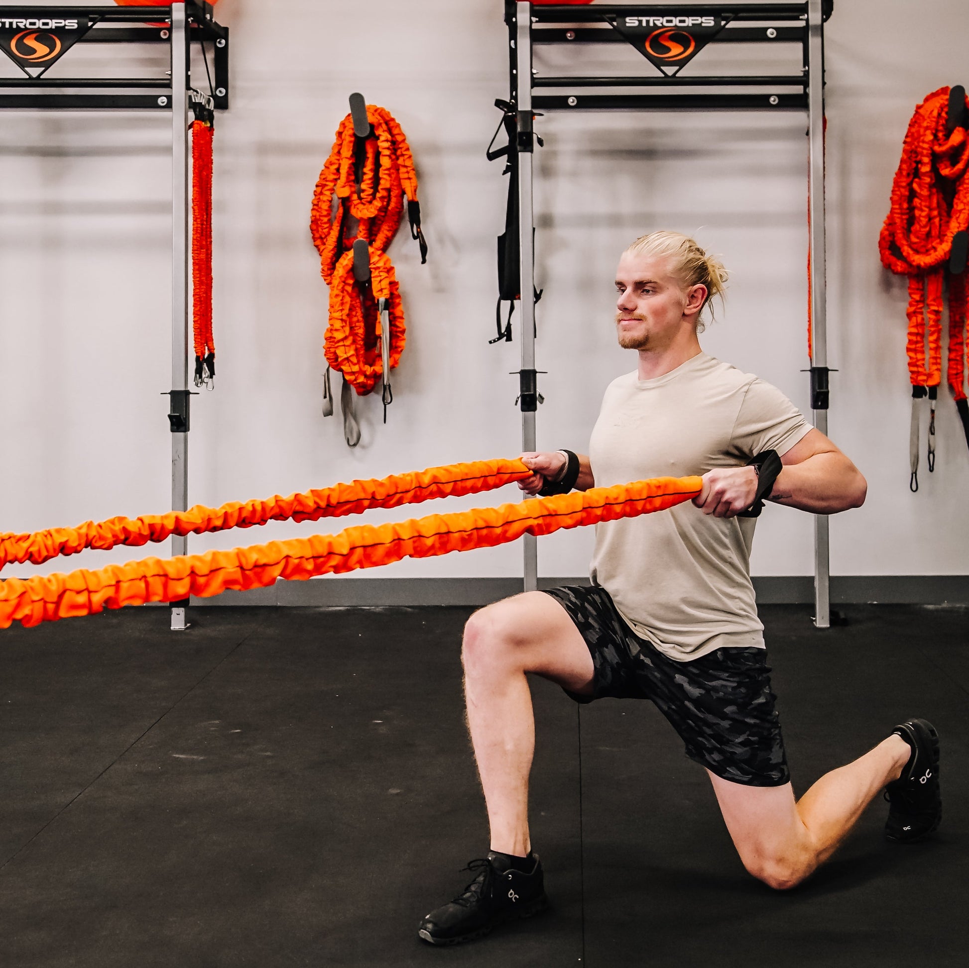 A man in a gym performing a lunge while holding a pair of bright orange battle ropes. The gym has black flooring, white walls, and various training equipment and orange ropes hanging on racks in the background.