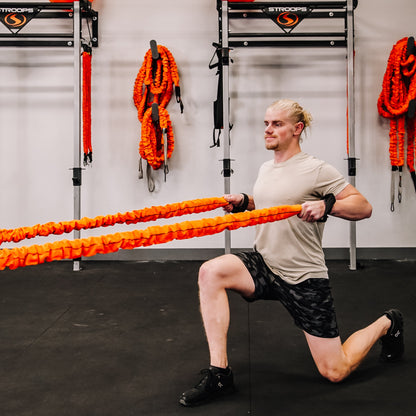 A man in a gym performing a lunge while holding a pair of bright orange battle ropes. The gym has black flooring, white walls, and various training equipment and orange ropes hanging on racks in the background.