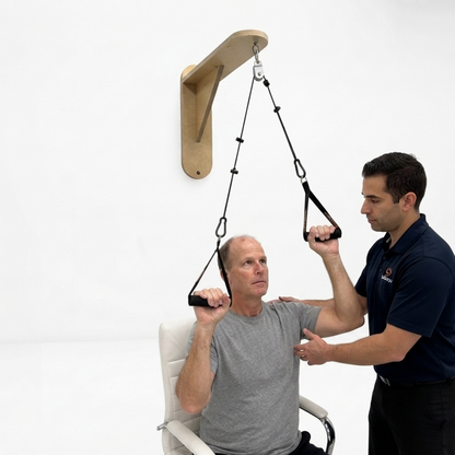 A man in a gray shirt sits in a chair using a wall-mounted resistance band with handles held by another man in a navy blue shirt, in a bright white studio setting.