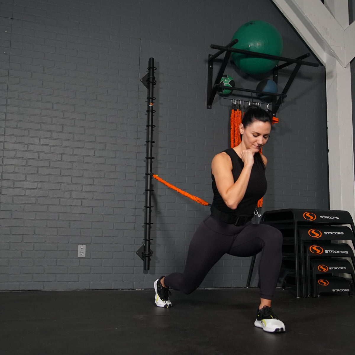 A woman in a black athletic outfit performs a lunge in a gym, using an orange resistance band attached to a wall rack. Black STROOPS step platforms are stacked on the floor to her right, and a green exercise ball is mounted on the wall above.