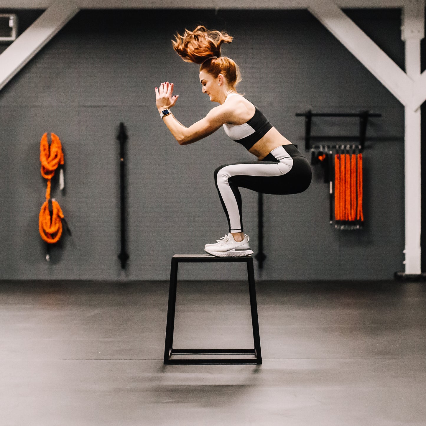 Woman performing a jump exercise on a black plyobox in a gym setting.