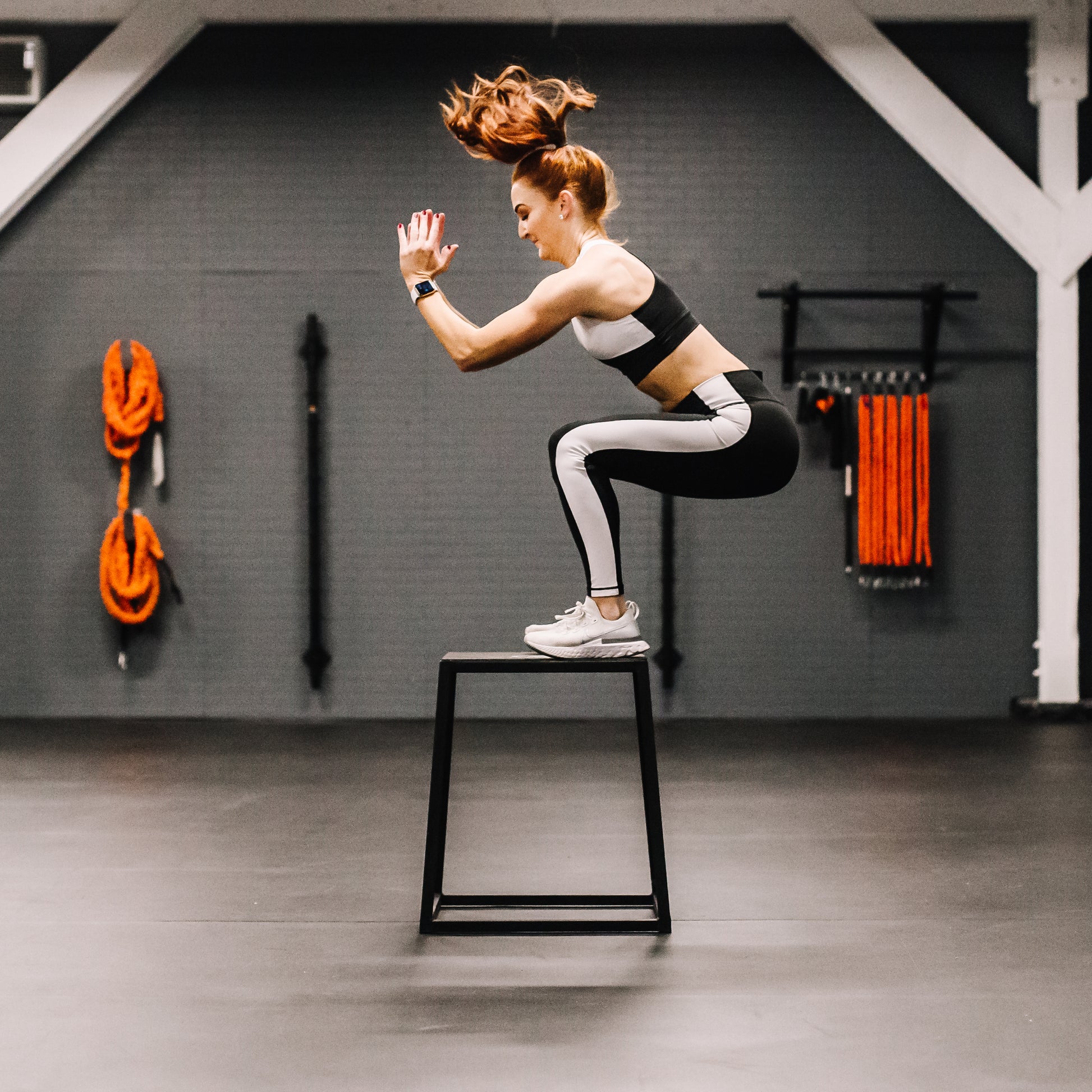 Woman performing a jump exercise on a black plyobox in a gym setting.
