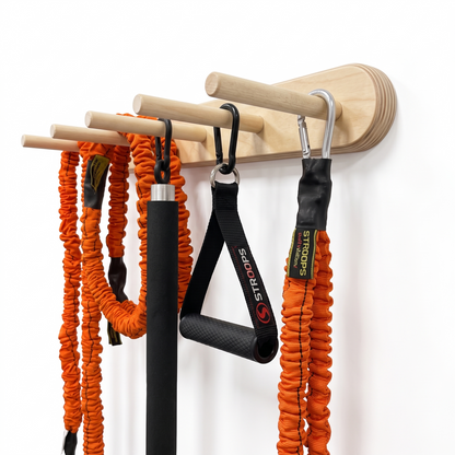 A wall-mounted wooden resistance tube holder with natural wood dowels, shown holding several fitness resistance tubes and black carabiners against a plain white wall.